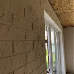 Wykończona elewacja budynku z imitacją cegły, widoczna z bliska pod kątem, z oknem przesuwnym i fragmentem drewnianej konstrukcji dachu.