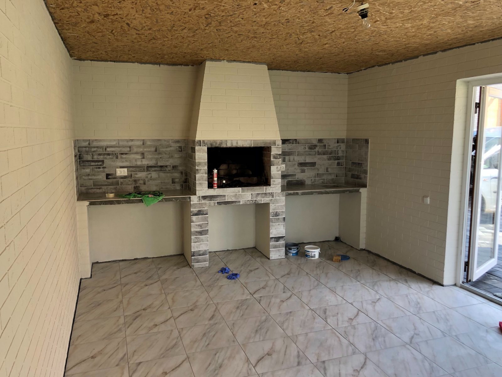 Wykończone wnętrze z ceglanym kominkiem i blatem roboczym, płytki podłogowe imitujące marmur, widoczne narzędzia i materiały budowlane.