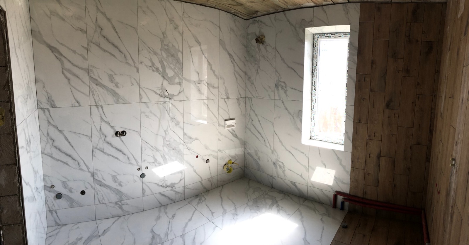 Wnętrze łazienki w trakcie remontu, ściany i podłoga wyłożone białymi płytkami imitującymi marmur, fragment ściany wykończony panelami drewnopodobnymi, widoczne instalacje hydrauliczne i okno.