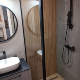 Natalia Maksymchak Adrian Józefowicz - Nowoczesna łazienka z kabiną prysznicową typu walk-in, drewnopodobnymi płytkami i okrągłym lustrem. Umywalka nablatowa na szafce z szufladami.