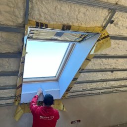 Natalia Maksymchak Adrian Józefowicz - Pracownik w czerwonej bluzie szpachluje płytę gipsową wokół okna dachowego na poddaszu z ociepleniem pianką PUR, widoczne metalowe profile konstrukcyjne.