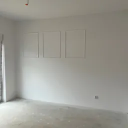 Jasne wnętrze po gładziowaniu, z dekoracyjnymi ramkami na ścianie i surową podłogą. Widoczne gniazdko elektryczne i wystający kabel z sufitu.