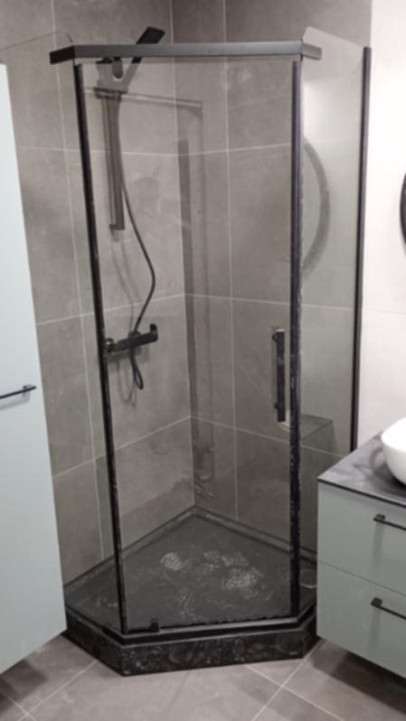 Nowoczesna kabina prysznicowa z czarnymi profilami i szklanymi ściankami, wkomponowana w szarą łazienkę z płytkami. Widoczna armatura i umywalka z szafką.