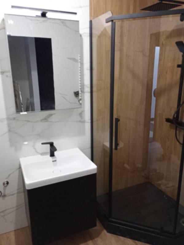 Nowoczesna łazienka z kabiną prysznicową w czarnej ramie, umywalką nablatową na szafce i lustrem z oświetleniem. Płytki imitujące marmur i drewno.