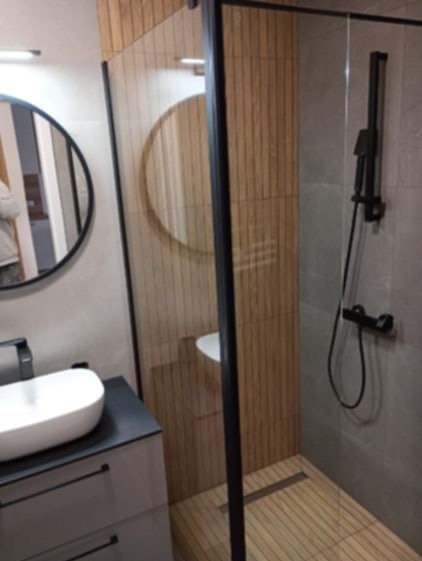 Nowoczesna łazienka z kabiną prysznicową typu walk-in, drewnopodobnymi płytkami i okrągłym lustrem. Umywalka nablatowa na szafce z szufladami.