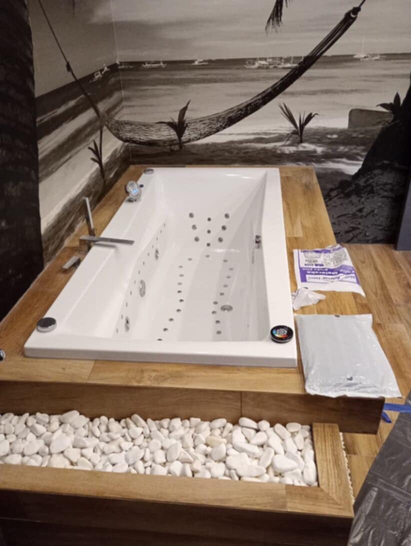 Biała wanna z hydromasażem, obudowana drewnem, z dekoracyjnymi białymi kamieniami. Na ścianie fototapeta z motywem plaży i hamaka. Częściowo widoczne materiały budowlane.