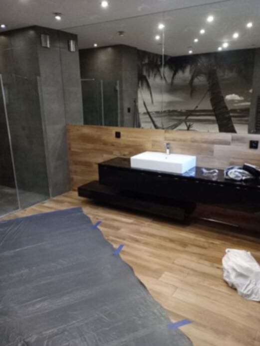 Nowoczesna łazienka z dużym lustrem, drewnianą boazerią i motywem tropikalnym na ścianie. Podłoga zabezpieczona folią malarską, biała umywalka na czarnej szafce.