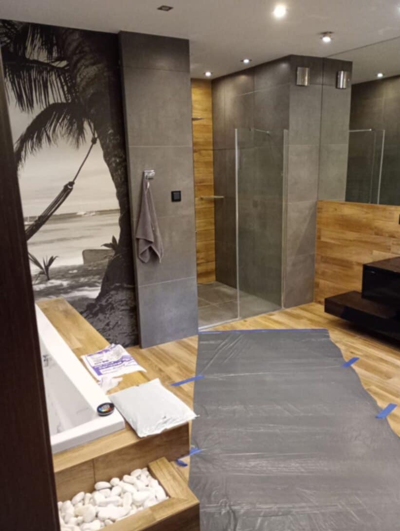 Nowoczesna łazienka w trakcie prac wykończeniowych: zabezpieczona podłoga, wanna z dekoracyjnymi kamieniami, motyw tropikalny na ścianie, kabina prysznicowa.