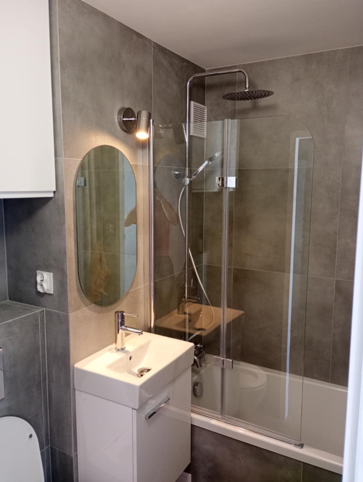 Nowoczesna łazienka z szarymi płytkami, owalnym lustrem z oświetleniem, białą umywalką z szafką i kabiną prysznicową nad wanną, tworząca spójną, minimalistyczną przestrzeń.