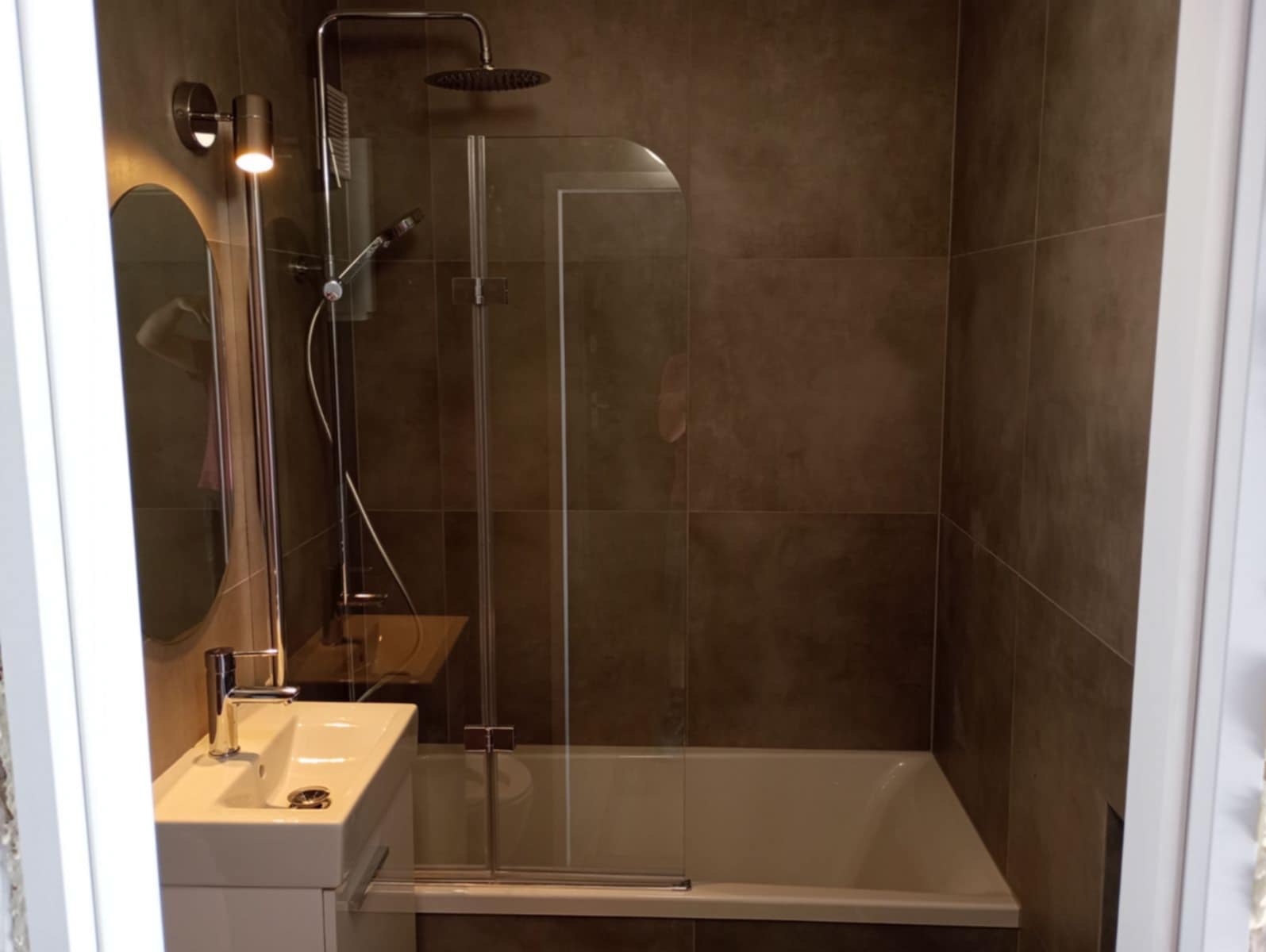 Nowoczesna łazienka z brązowymi płytkami, kabiną prysznicową z szybą i owalnym lustrem z podświetleniem LED. Minimalistyczna umywalka z szafką podwieszaną.