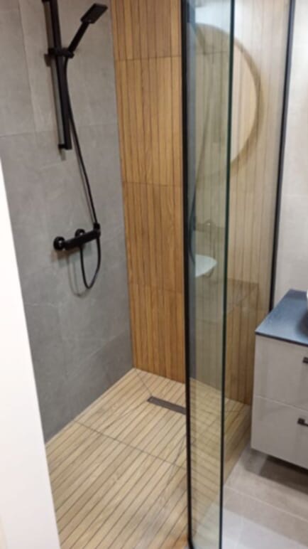Nowoczesna kabina prysznicowa z drewnianą podłogą i panelami ściennymi, czarna armatura, szklana ścianka działowa, szare płytki. Wykończenie łazienki w naturalnym stylu.