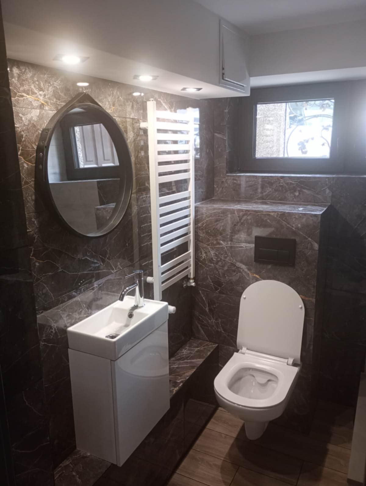 Elegancka łazienka z marmurowymi płytkami, owalnym lustrem, białą umywalką i grzejnikiem drabinkowym. Nowoczesny design i ciepłe oświetlenie tworzą przytulną atmosferę.