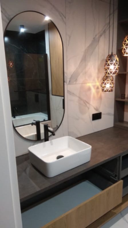 Nowoczesna łazienka z owalnym lustrem, białą umywalką nablatową i marmurową ścianą. Widoczne geometryczne lampy wiszące i drewniane wykończenia szafek.
