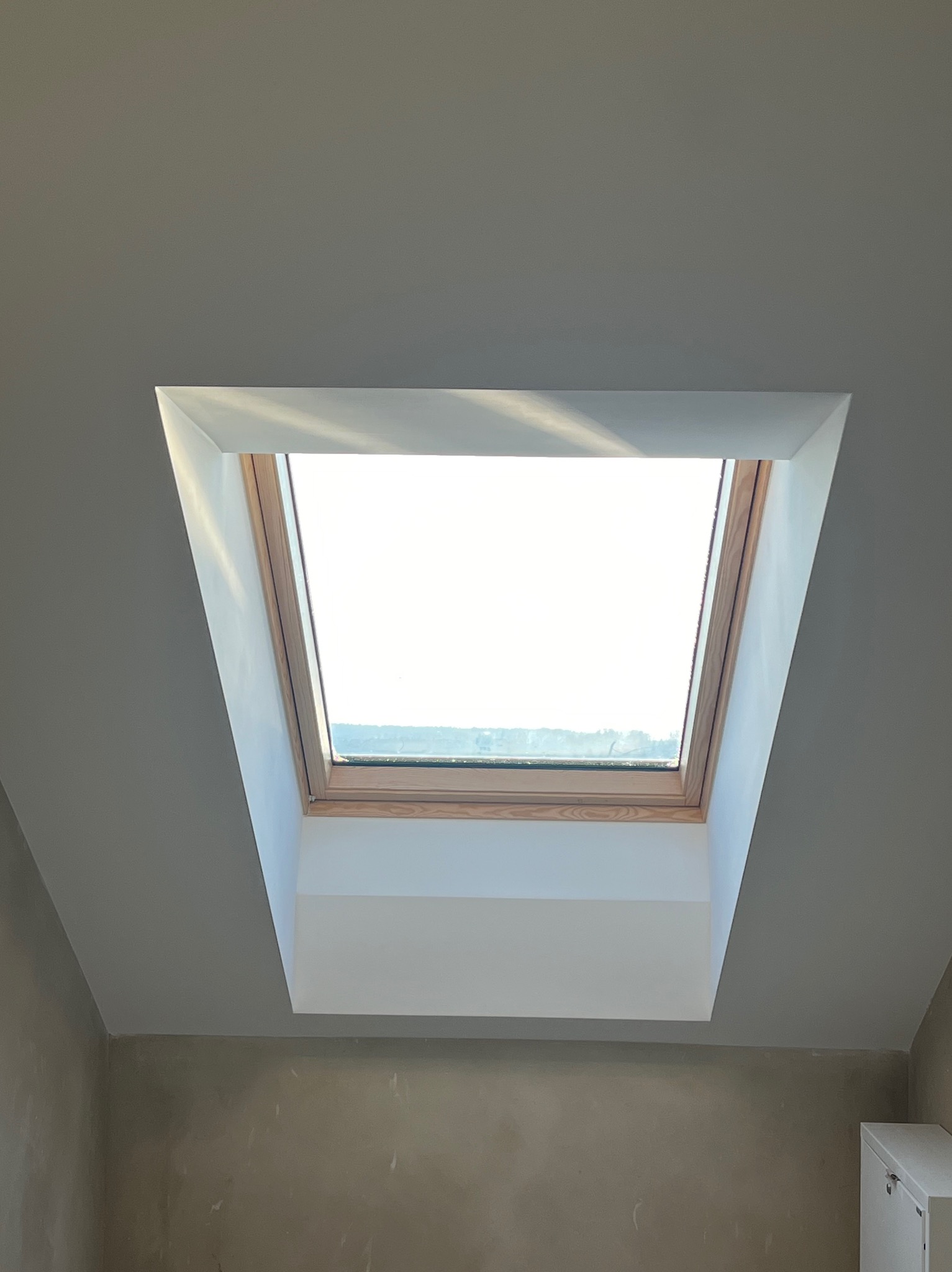 Wykończone okno dachowe z drewnianą ramą i białymi obramowaniami, wkomponowane w szary sufit poddasza. Świeże gładzie na ścianach. Widok na zewnątrz przez okno.