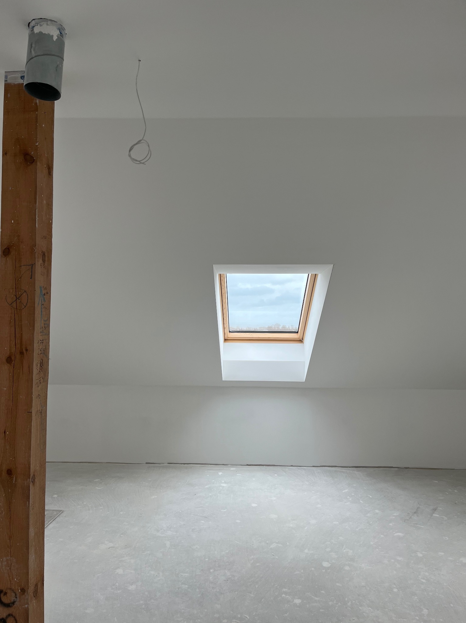 Poddasze ze świeżo wygładzonymi ścianami i oknem dachowym. Widoczna drewniana belka z oznaczeniami oraz wystający przewód elektryczny.