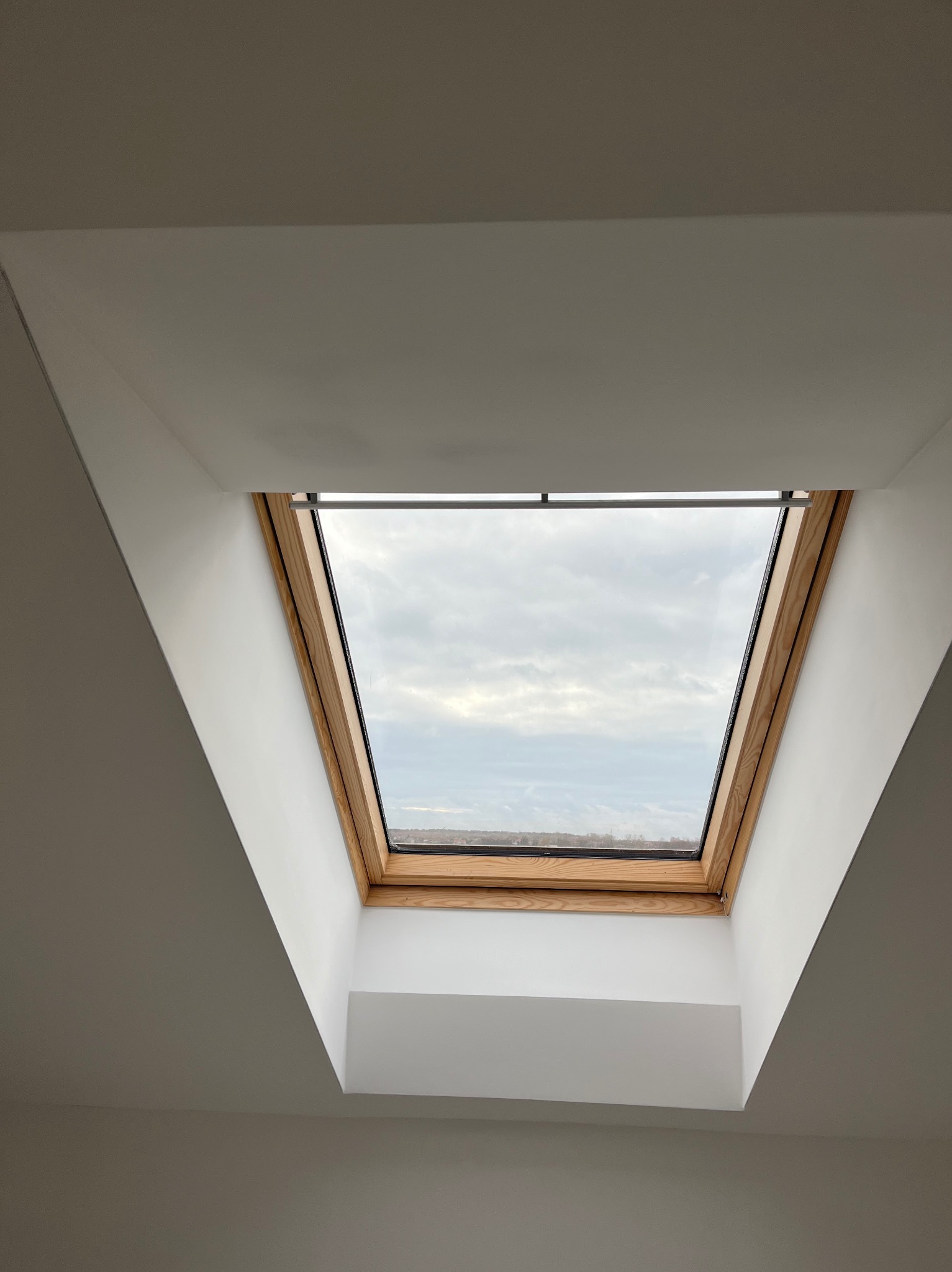 Okno dachowe z drewnianą ramą, wpuszczone w biały sufit, widok na pochmurne niebo i odległy krajobraz. Minimalistyczne wykończenie wnętrza.