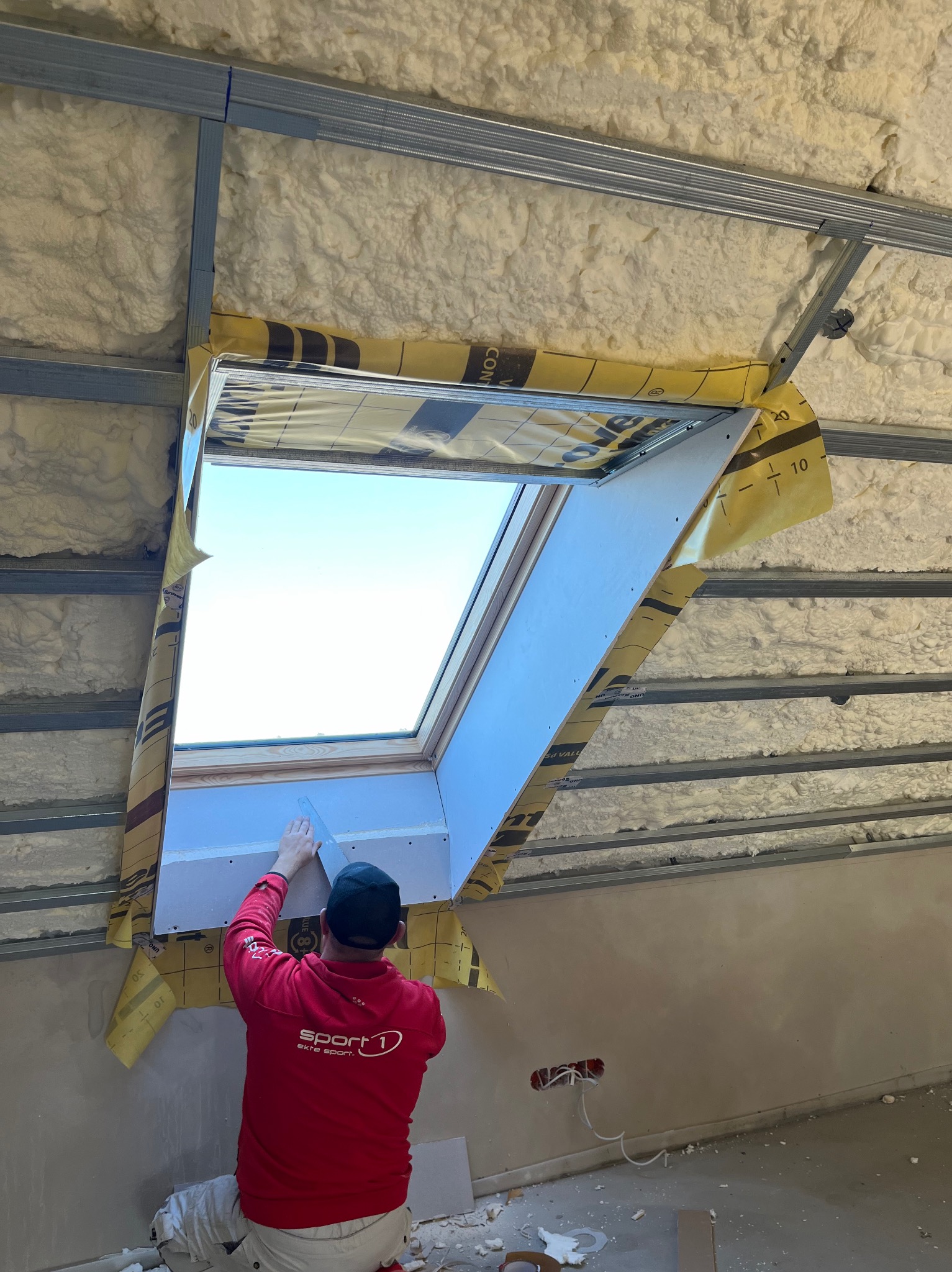 Pracownik w czerwonej bluzie szpachluje płytę gipsową wokół okna dachowego na poddaszu z ociepleniem pianką PUR, widoczne metalowe profile konstrukcyjne.