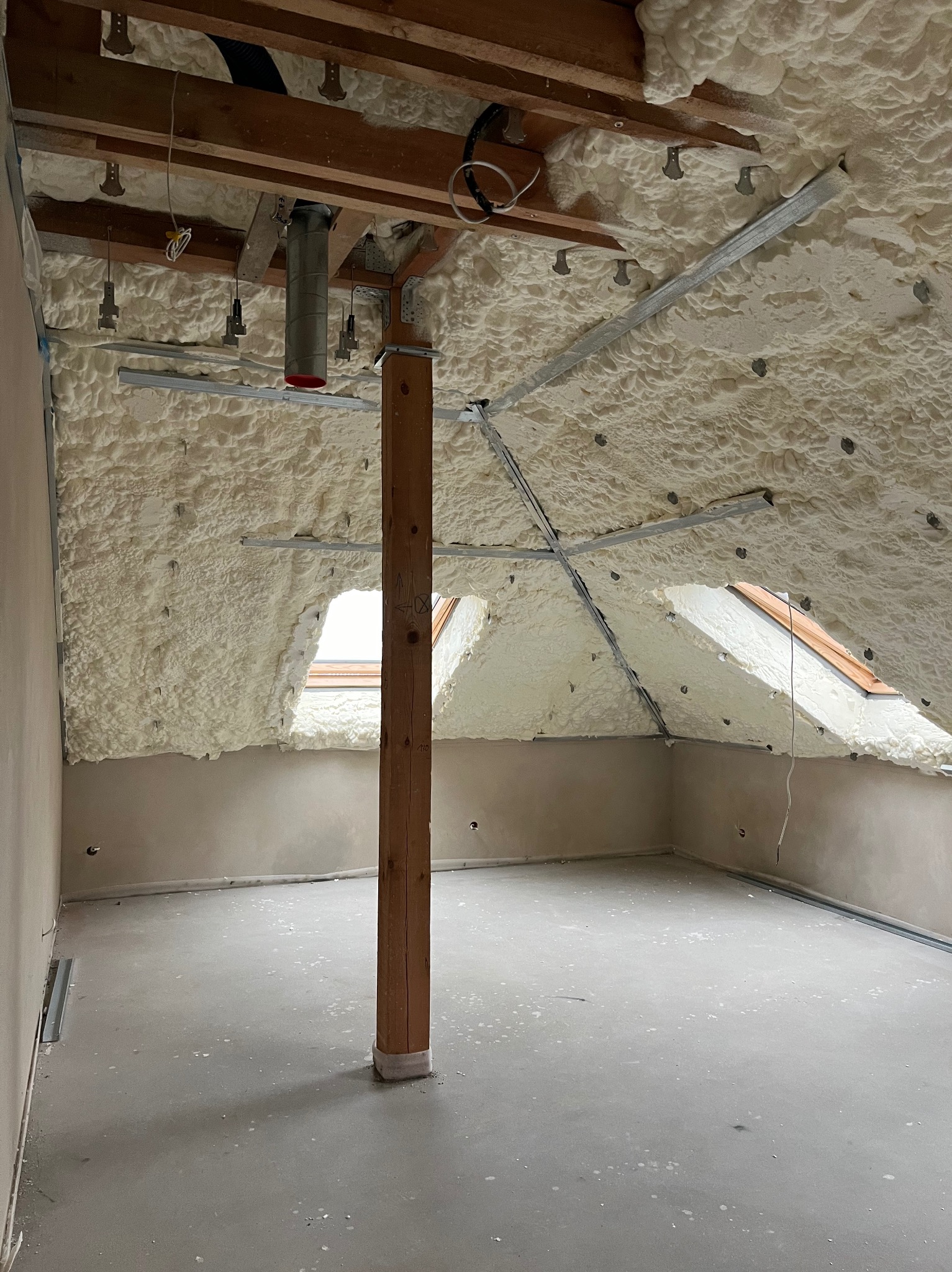 Poddasze po izolacji pianą PUR: biała piana pokrywa skośny sufit z oknami dachowymi, widoczne drewniane belki i metalowe profile, surowy beton na podłodze.