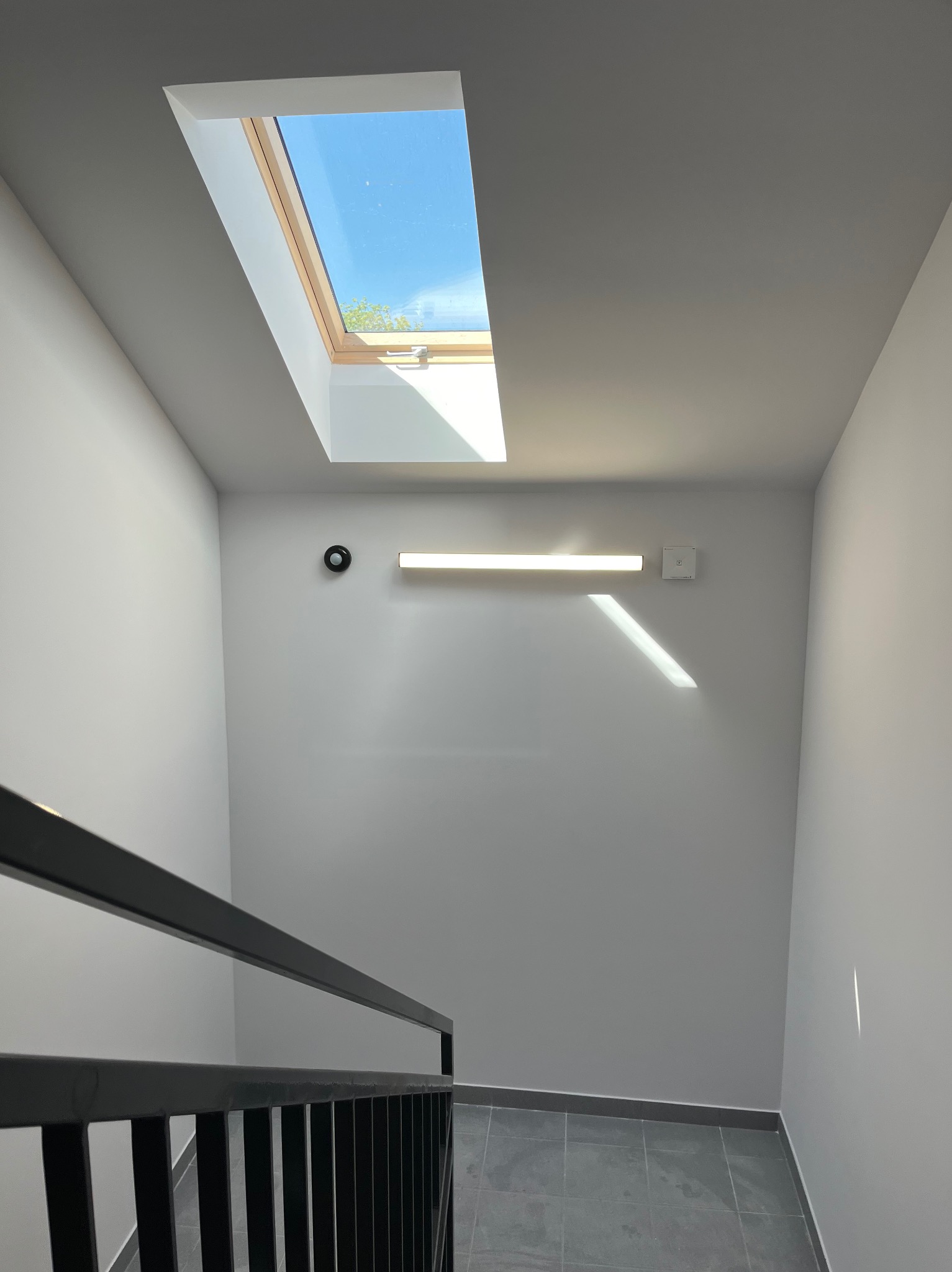 Klatka schodowa z szarą posadzką i czarną balustradą. W górze okno dachowe wpuszcza światło. Na ścianie lampa i czujnik dymu. Minimalistyczny design.