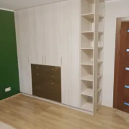 Biała szafa wnękowa z drewnianymi frontami, brązowe szuflady i otwarty regał z p&oacute;łkami, widok na podłogę z paneli.