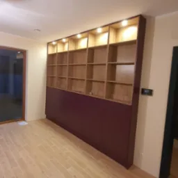 Wysoka zabudowa ścienna z otwartymi, podświetlanymi półkami z drewna i dolnymi szafkami w kolorze burgundu, na podłodze płytki imitujące drewno.