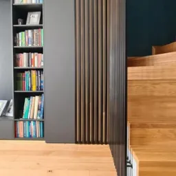 Nowoczesne wnętrze w Koszalinie: drewniana podłoga, schody z jasnego drewna i regał z książkami obok ażurowej ścianki działowej z pionowych listew.