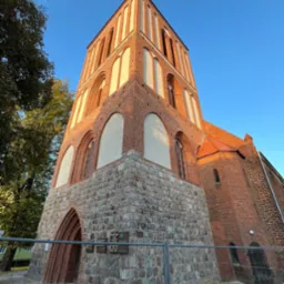 Gotycki kościół z czerwonej cegły i kamienną podstawą, sfotografowany z dołu pod kątem, z widocznymi tablicami pamiątkowymi i fragmentem ogrodzenia.