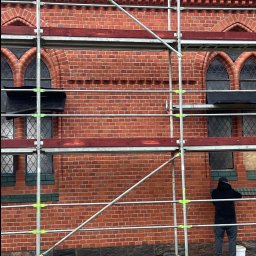 JAROSŁAW KACZMAREK KLINKIER-FUG - Ceglana elewacja budynku w rusztowaniach, widoczne zdobienia i okna. Osoba w ciemnej bluzie stoi obok rusztowania. Prace renowacyjne w toku, widoczny fragment dachu.