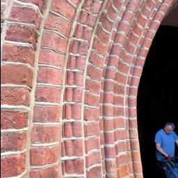 JAROSŁAW KACZMAREK KLINKIER-FUG - Ceglany łuk z wyraźnie odnowioną spoiną, fragment muru z bliska. W tle sylwetka mężczyzny w niebieskiej koszulce, prawdopodobnie podczas pracy.