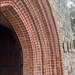 JAROSŁAW KACZMAREK KLINKIER-FUG - Ceglany łuk nad wejściem do zabytkowej kamienicy, z drewnianymi drzwiami i zdobieniami z kutego żelaza. Część kamiennej elewacji z tablicami pamiątkowymi.