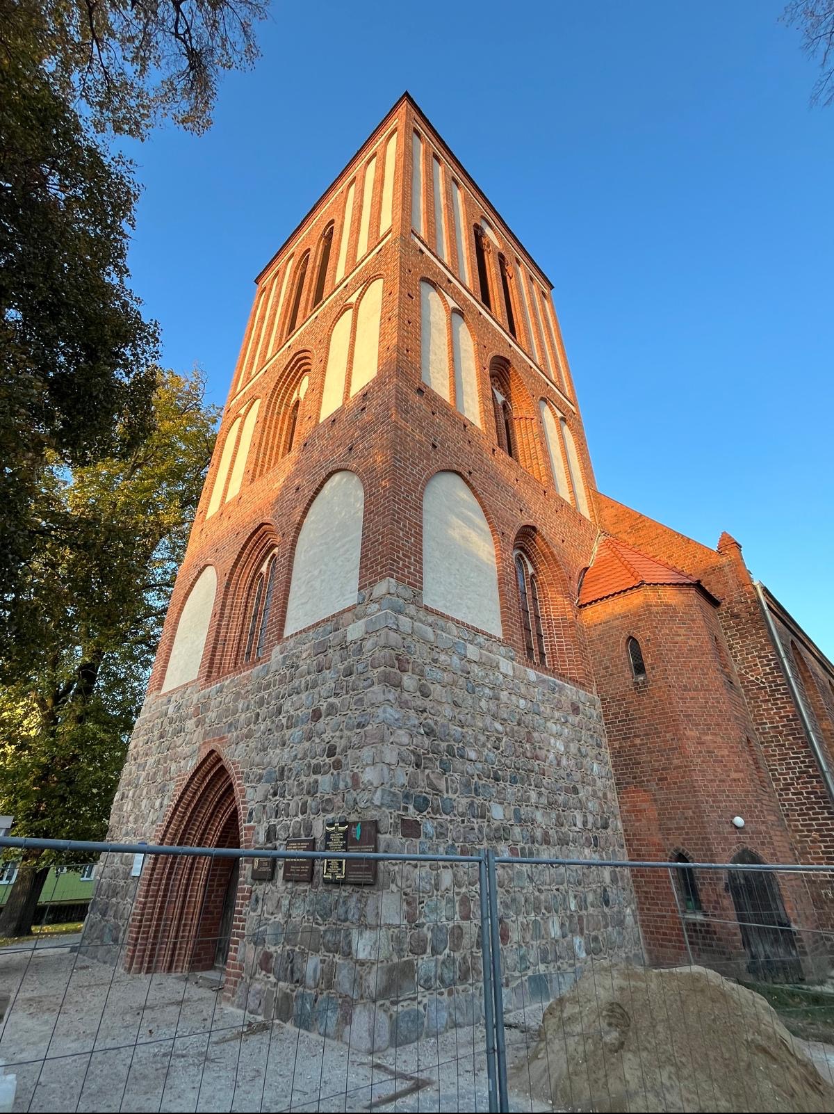 Gotycki kościół z czerwonej cegły i kamienną podstawą, sfotografowany z dołu pod kątem, z widocznymi tablicami pamiątkowymi i fragmentem ogrodzenia.