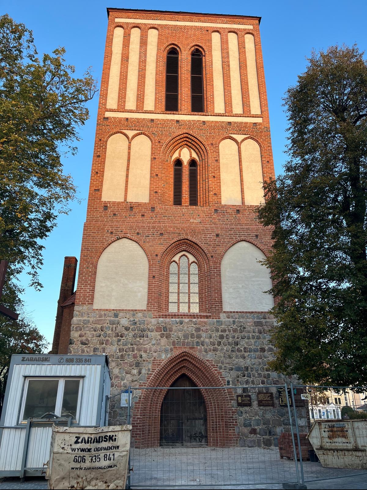 Ceglana wieża kościoła z elementami kamiennymi, widoczna z dołu, otoczona drzewami. Na pierwszym planie kontener budowlany i ogrodzenie.