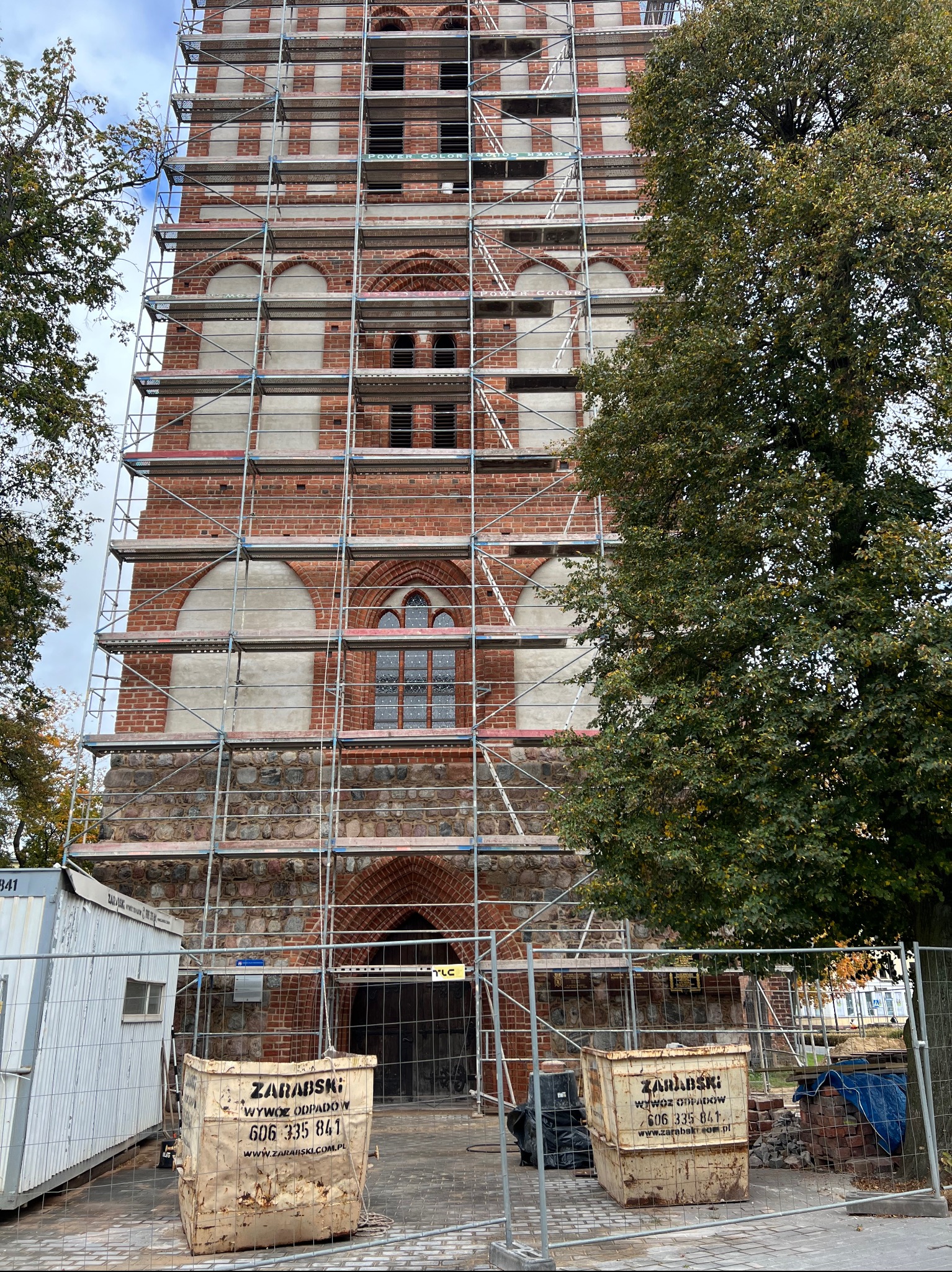 Wysoka wieża kościoła w rusztowaniach, z widocznymi detalami architektonicznymi i elementami renowacji. Dwa kontenery na odpady przed wejściem. Częściowo zasłonięta przez drzewo.