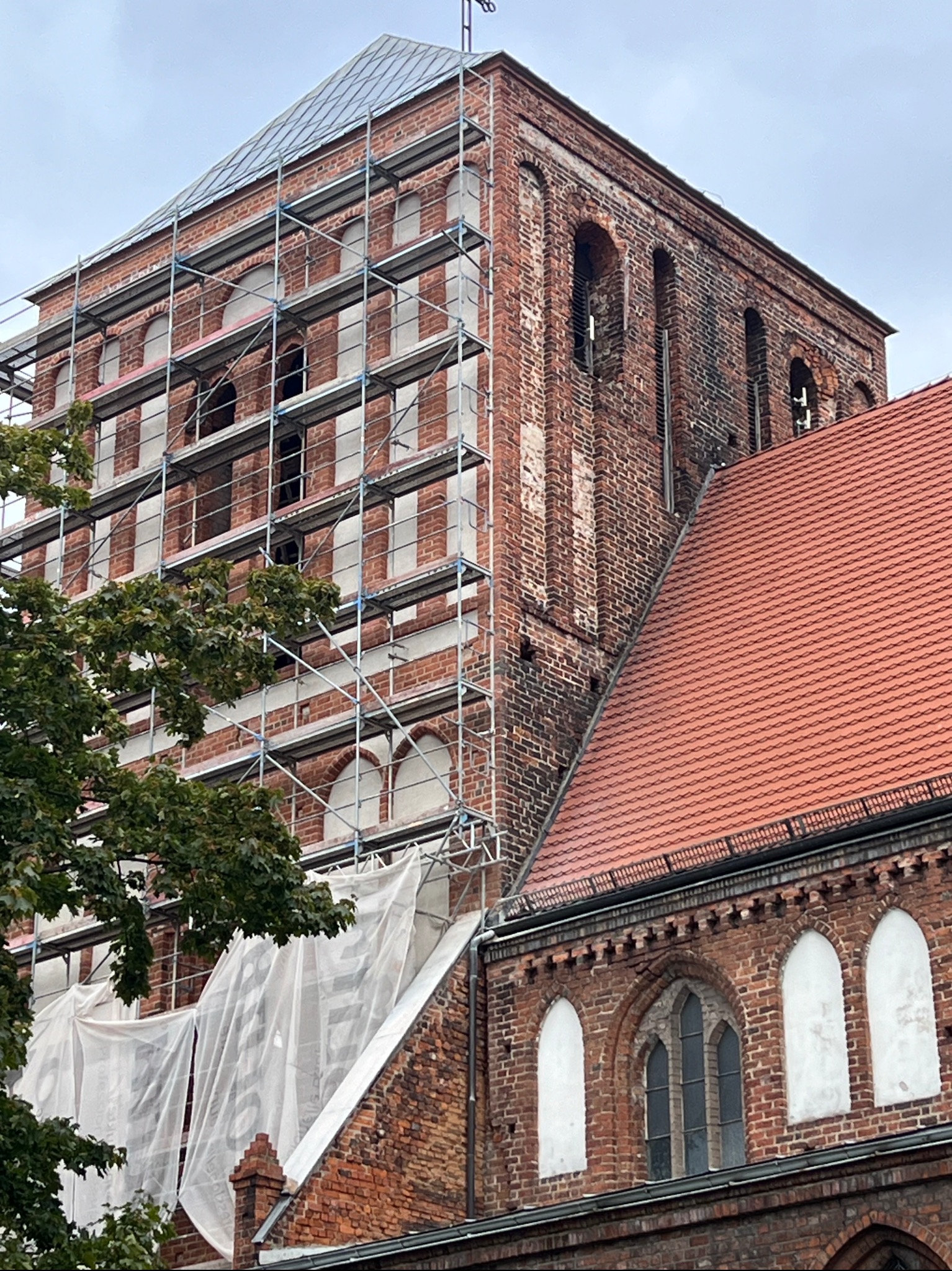 Ceglana wieża kościoła w rusztowaniach, częściowo zakryta płachtą ochronną. Widoczny fragment dachu z czerwonej dachówki. Prace renowacyjne w toku.