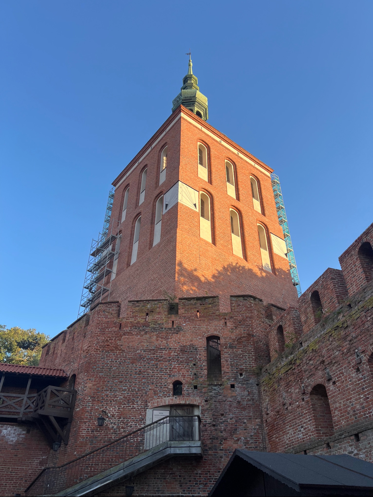 Ceglana wieża z rusztowaniem w słoneczny dzień. Widoczne okna z łukami i zielona iglica. Fragment muru obronnego.