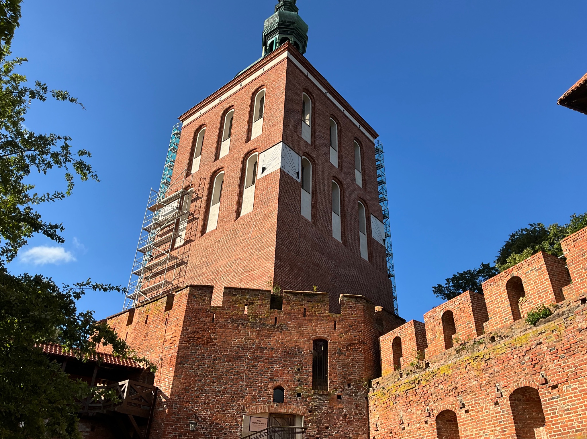 Kcynia: Ceglana wieża z zegarem słonecznym w trakcie renowacji. Widoczne rusztowania i fragment muru obronnego. Błękitne niebo w tle. Prace konserwatorskie.