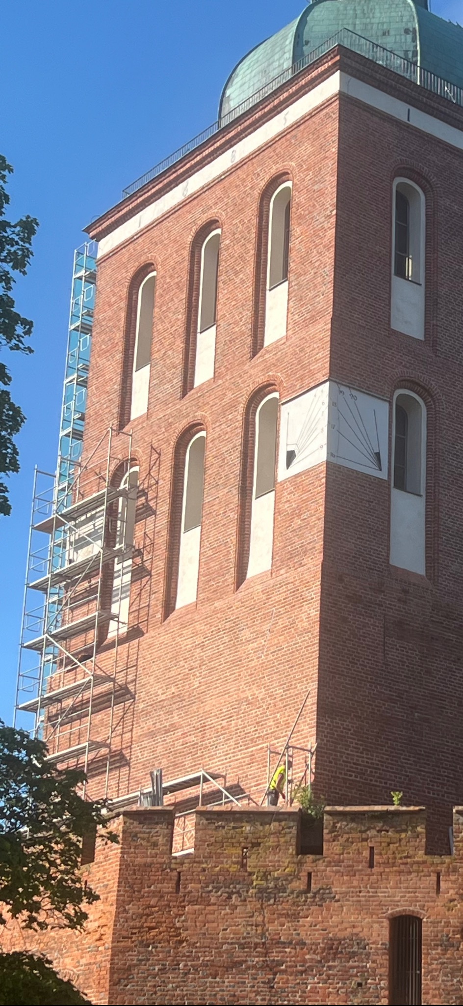 Ceglana wieża z zegarem słonecznym w trakcie renowacji; widoczne rusztowania i pracownik w odblaskowej kamizelce. Błękitne niebo w tle.