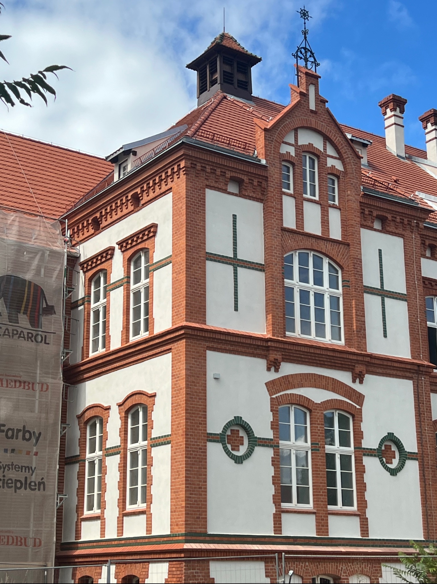 Odrestaurowana elewacja kamienicy z czerwonej cegły i białego tynku. Widoczne detale architektoniczne, okna z białymi ramami oraz rusztowania z siatką ochronną.