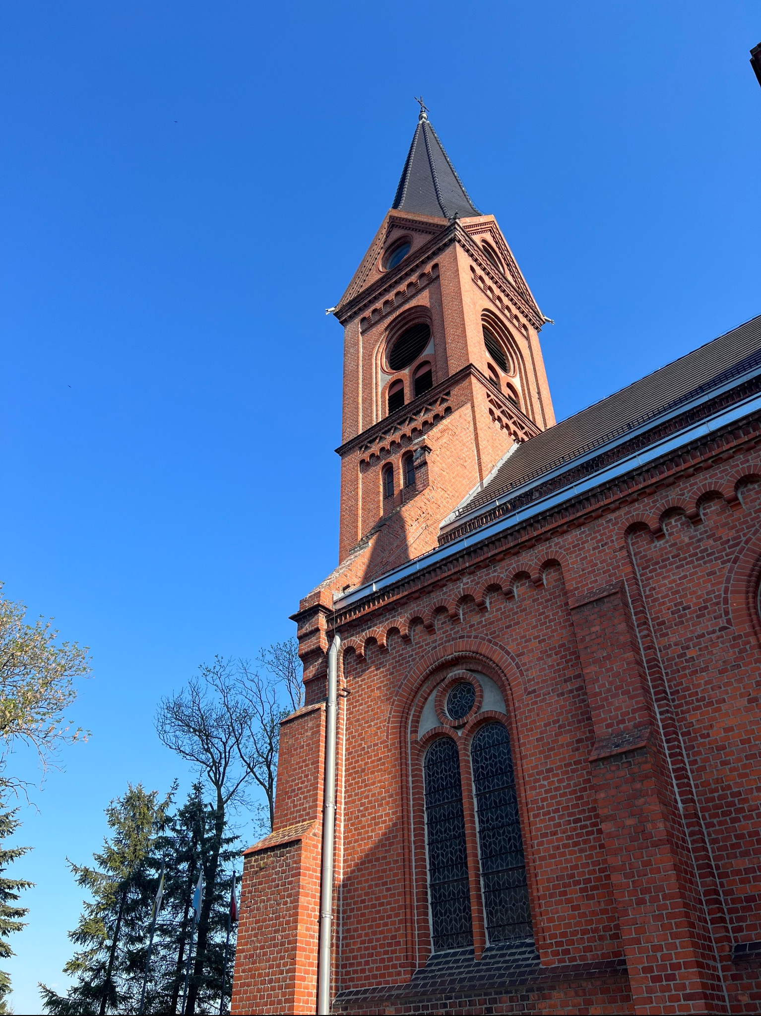 Ceglana wieża kościelna z czarnym dachem na tle błękitnego nieba, widziana z dołu. Architektura sakralna z detalami z cegły.