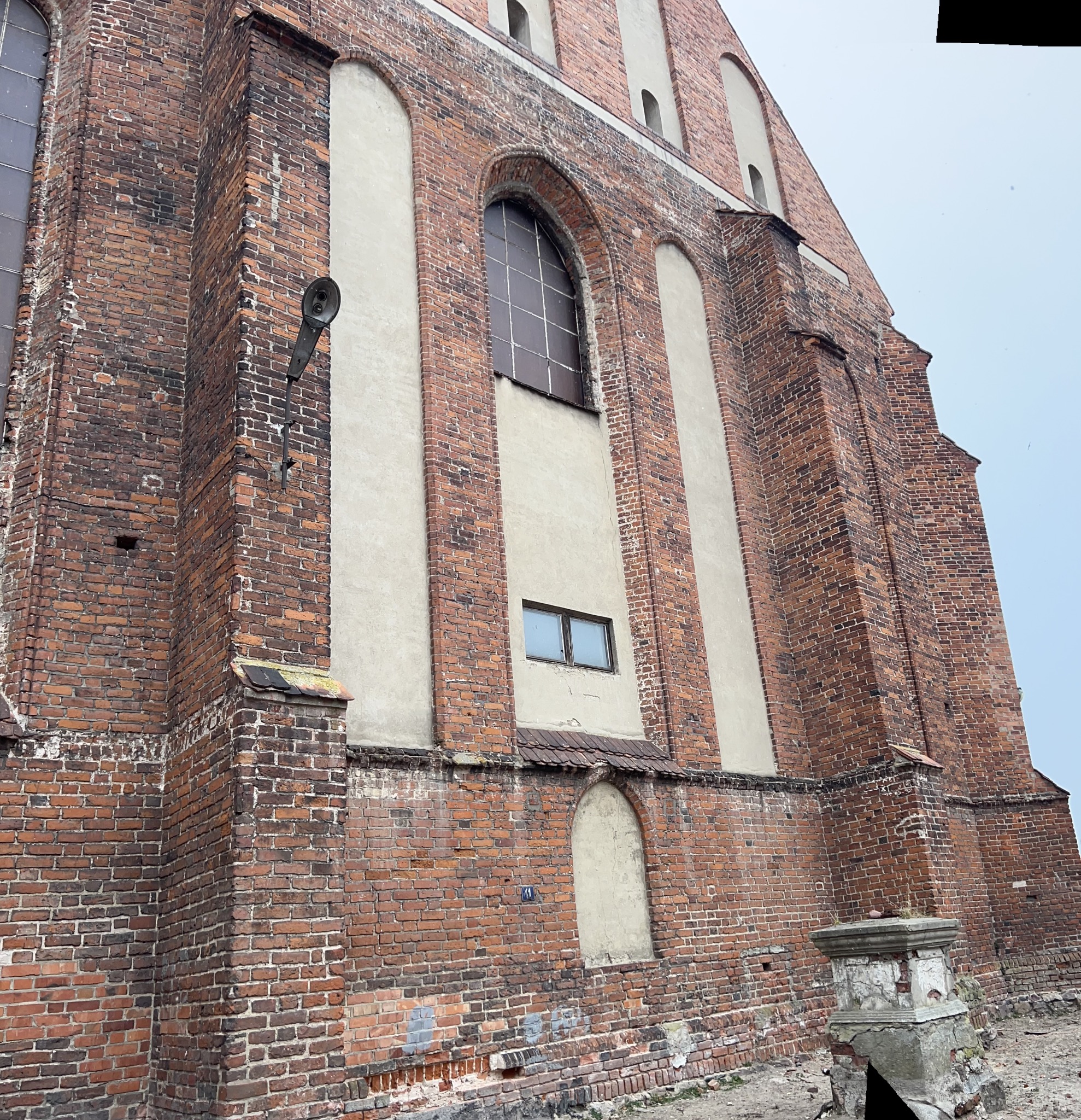 Ceglana fasada gotyckiego kościoła z łukowymi oknami i betonowymi wstawkami, widoczny fragment zniszczonego postumentu i lampa na ścianie.