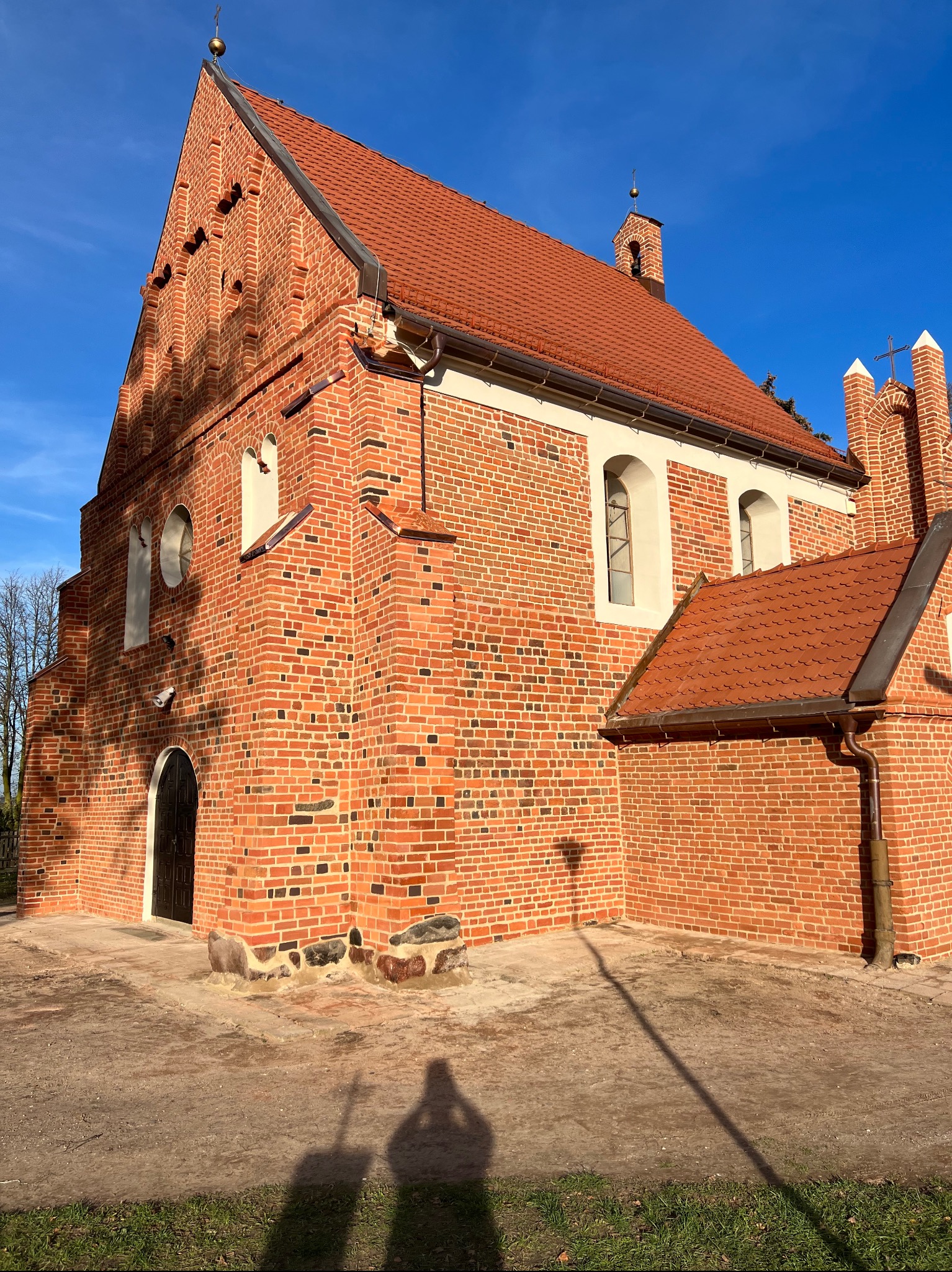 Ceglany kościół z czerwoną dachówką, widok z dołu, cień osoby na pierwszym planie. Architektura sakralna po renowacji elewacji.