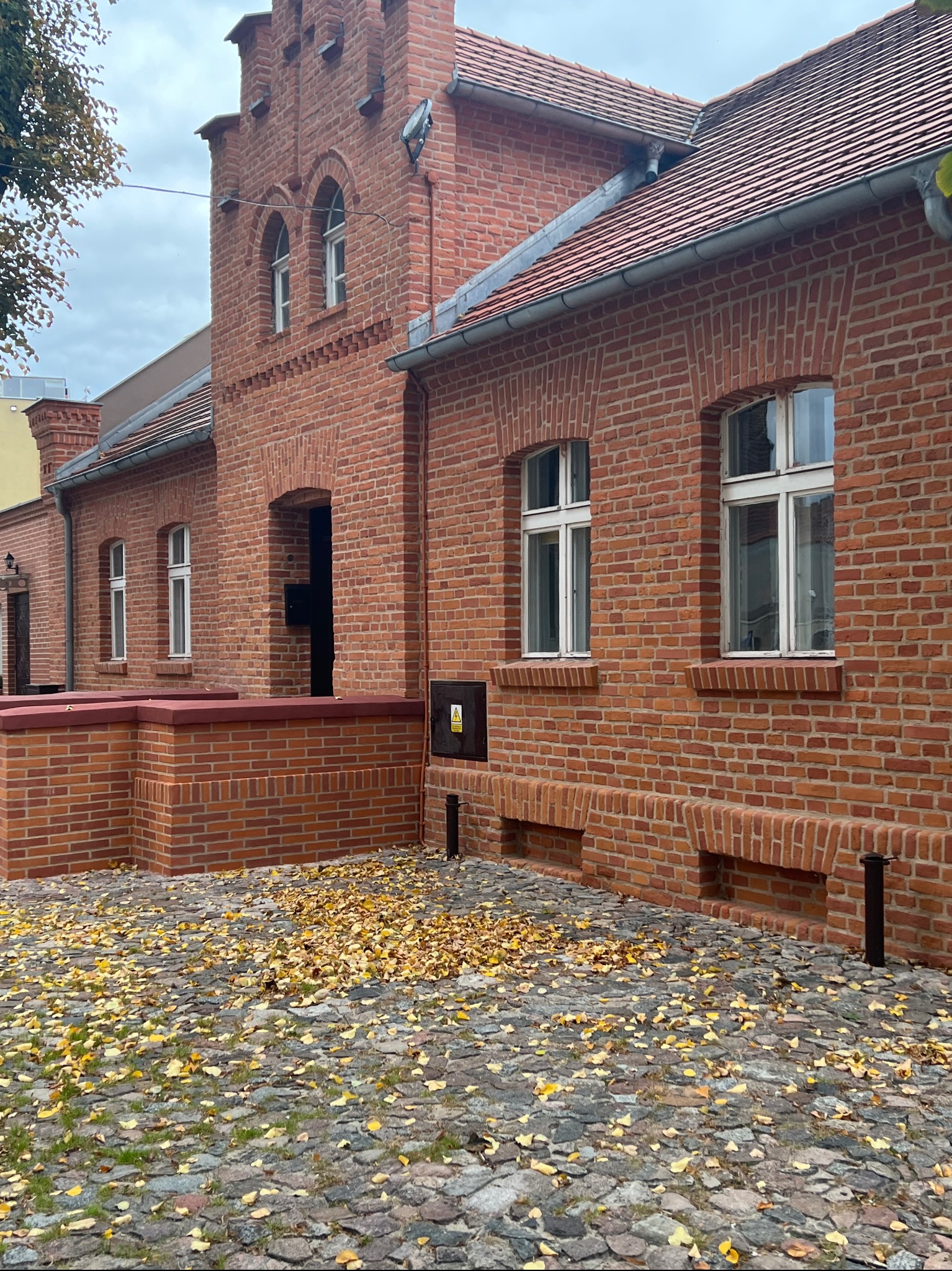 Ceglana elewacja odnowionego budynku z białymi oknami i brukowanym chodnikiem pokrytym jesiennymi liśćmi. Widoczne detale architektoniczne.