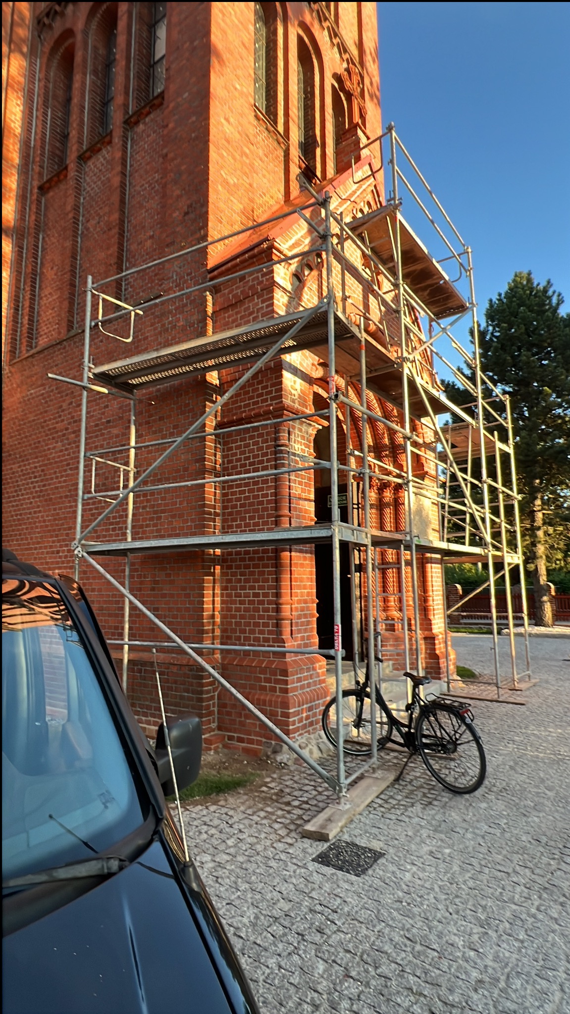 Ceglana wieża kościoła w rusztowaniach, rower oparty o ścianę, fragment auta w kadrze. Renowacja elewacji w toku, ciepłe światło poranka.