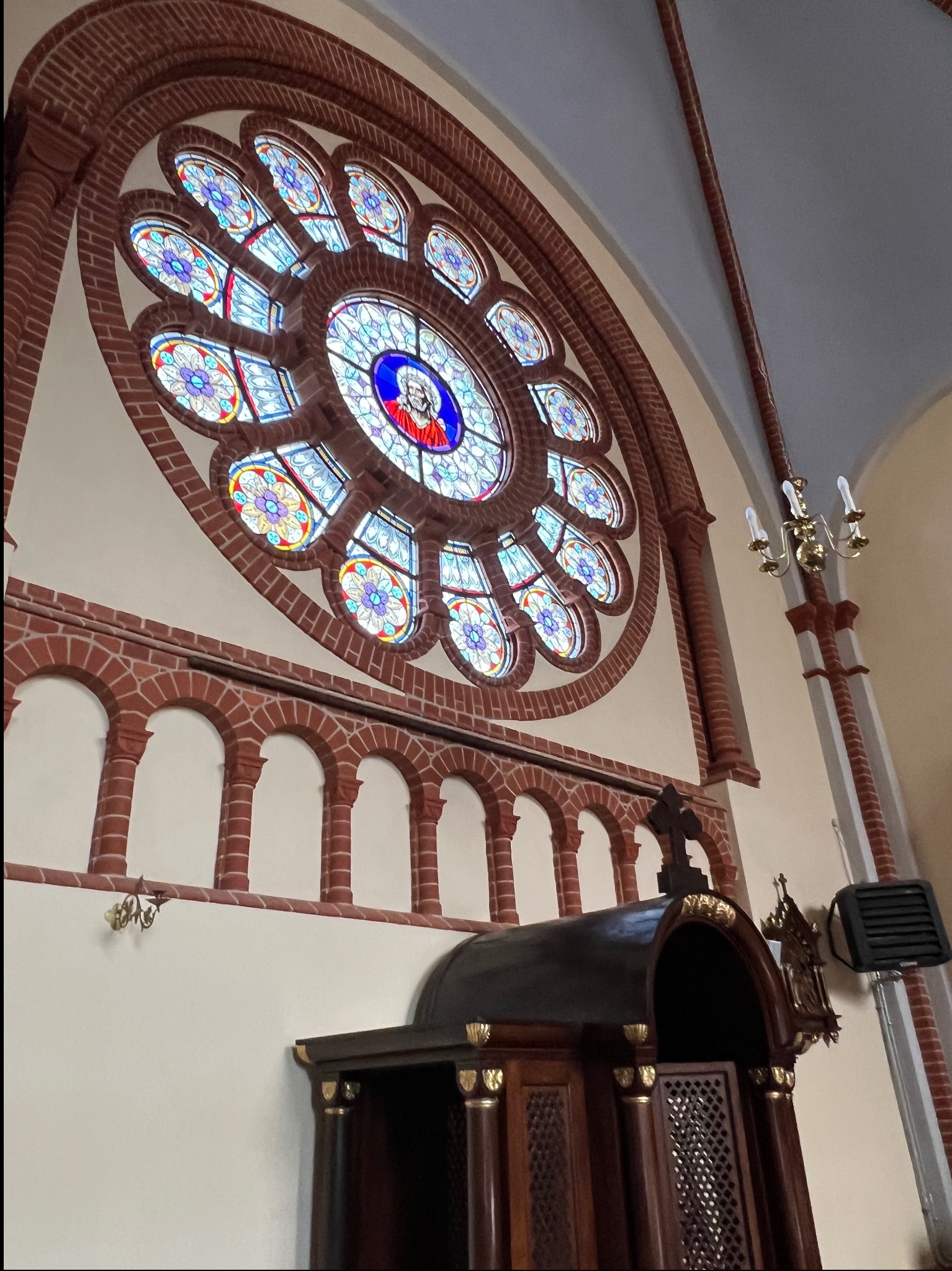 Wnętrze kościoła z dużym, okrągłym witrażem w rozecie z ceglaną obudową. Na dole widoczny drewniany konfesjonał z dekoracyjnymi elementami.