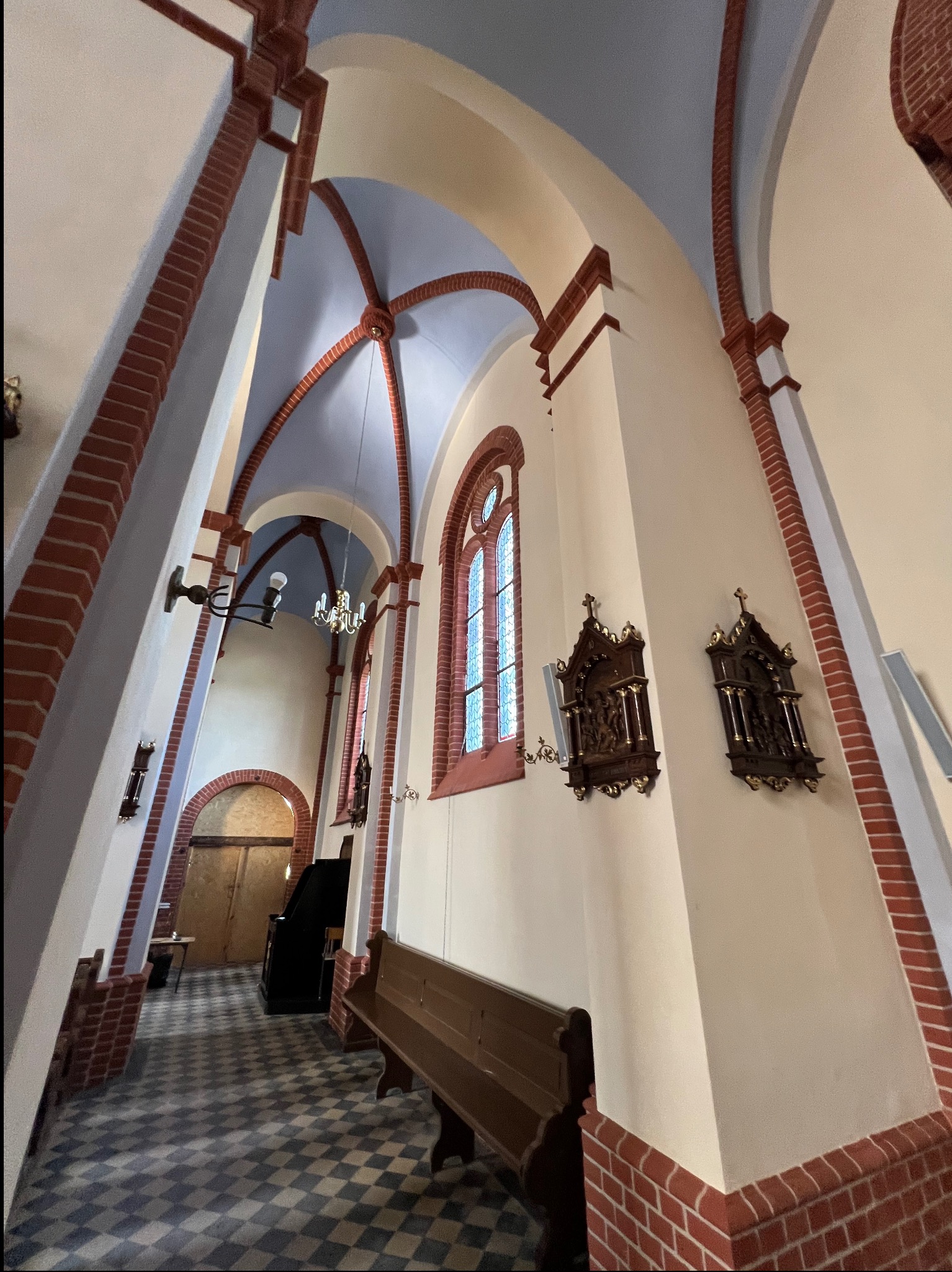 Wnętrze kościoła z łukami, ceglanymi detalami i drewnianymi rzeźbami na ścianach. Posadzka w szachownicę, ławki i oświetlenie podkreślają historyczny charakter.