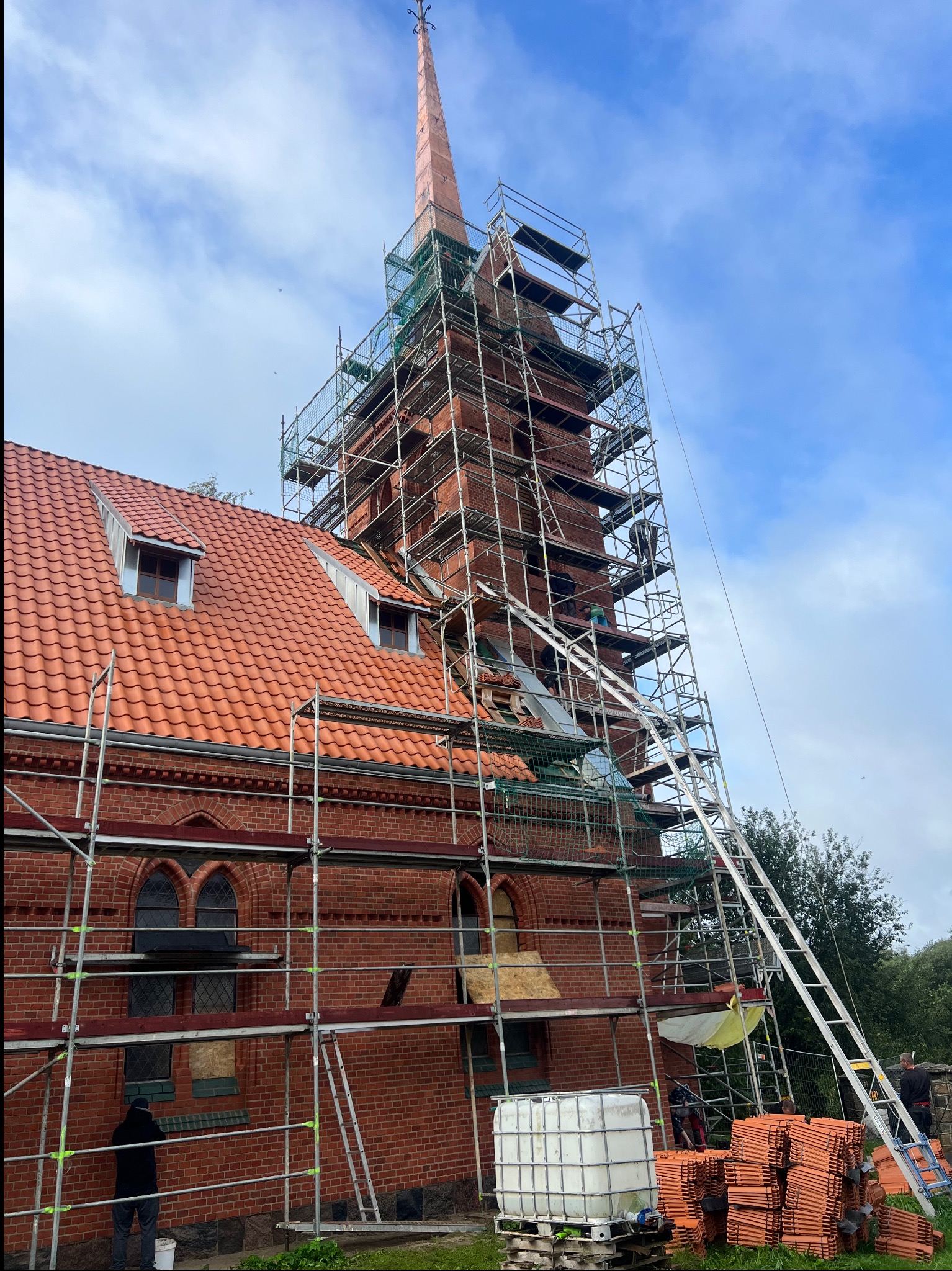 Ceglany kościół w rusztowaniach podczas renowacji, widoczne nowe dachówki, drabina oparta o rusztowanie, częściowo zasłonięte okna.