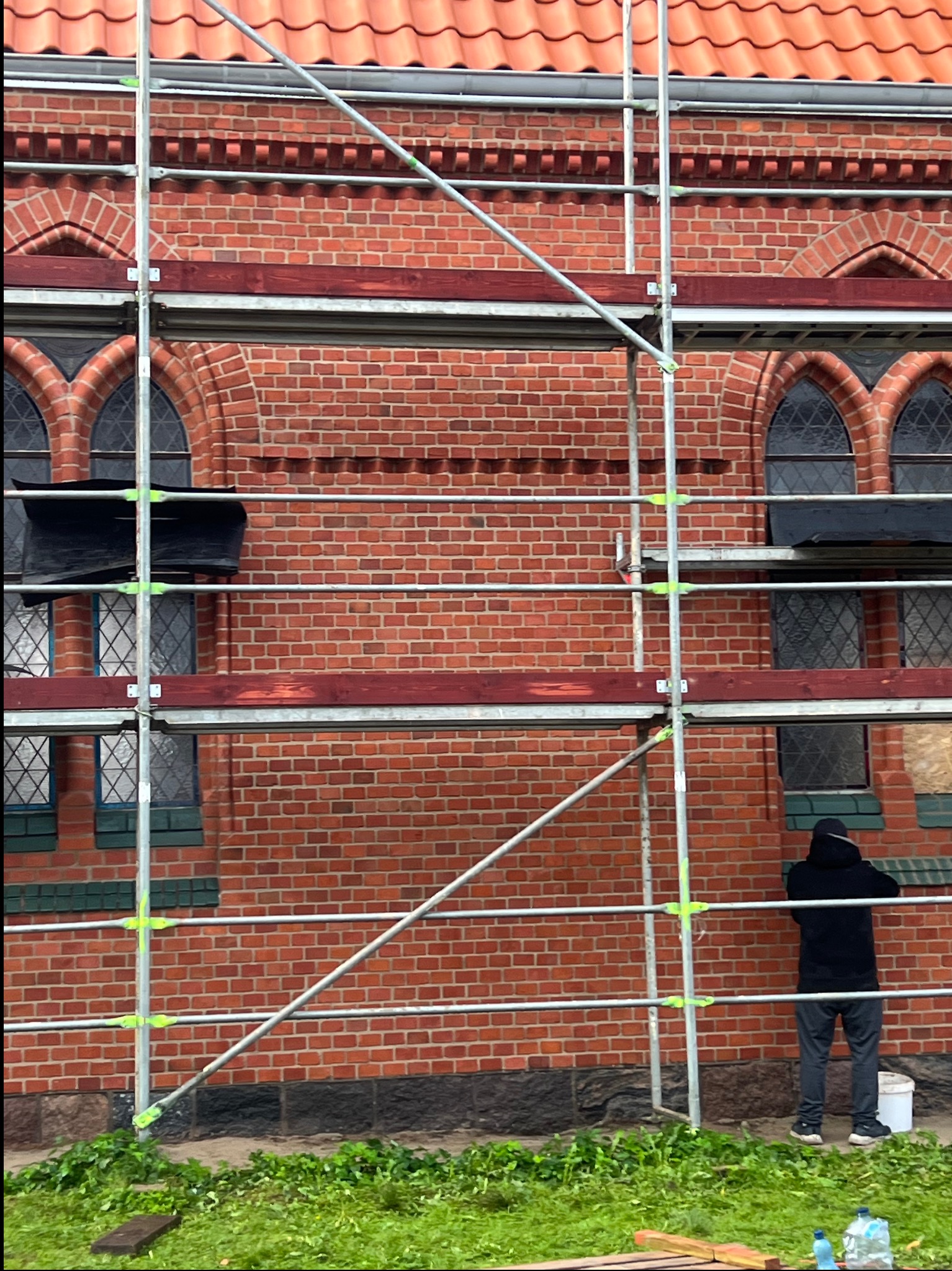 Ceglana elewacja budynku w rusztowaniach, widoczne zdobienia i okna. Osoba w ciemnej bluzie stoi obok rusztowania. Prace renowacyjne w toku, widoczny fragment dachu.