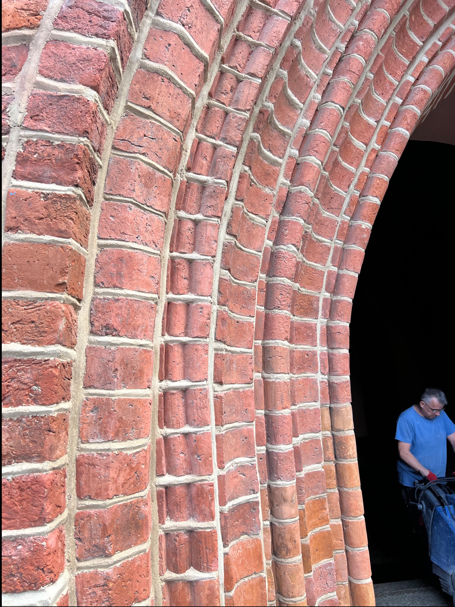 Ceglany łuk z wyraźnie odnowioną spoiną, fragment muru z bliska. W tle sylwetka mężczyzny w niebieskiej koszulce, prawdopodobnie podczas pracy.