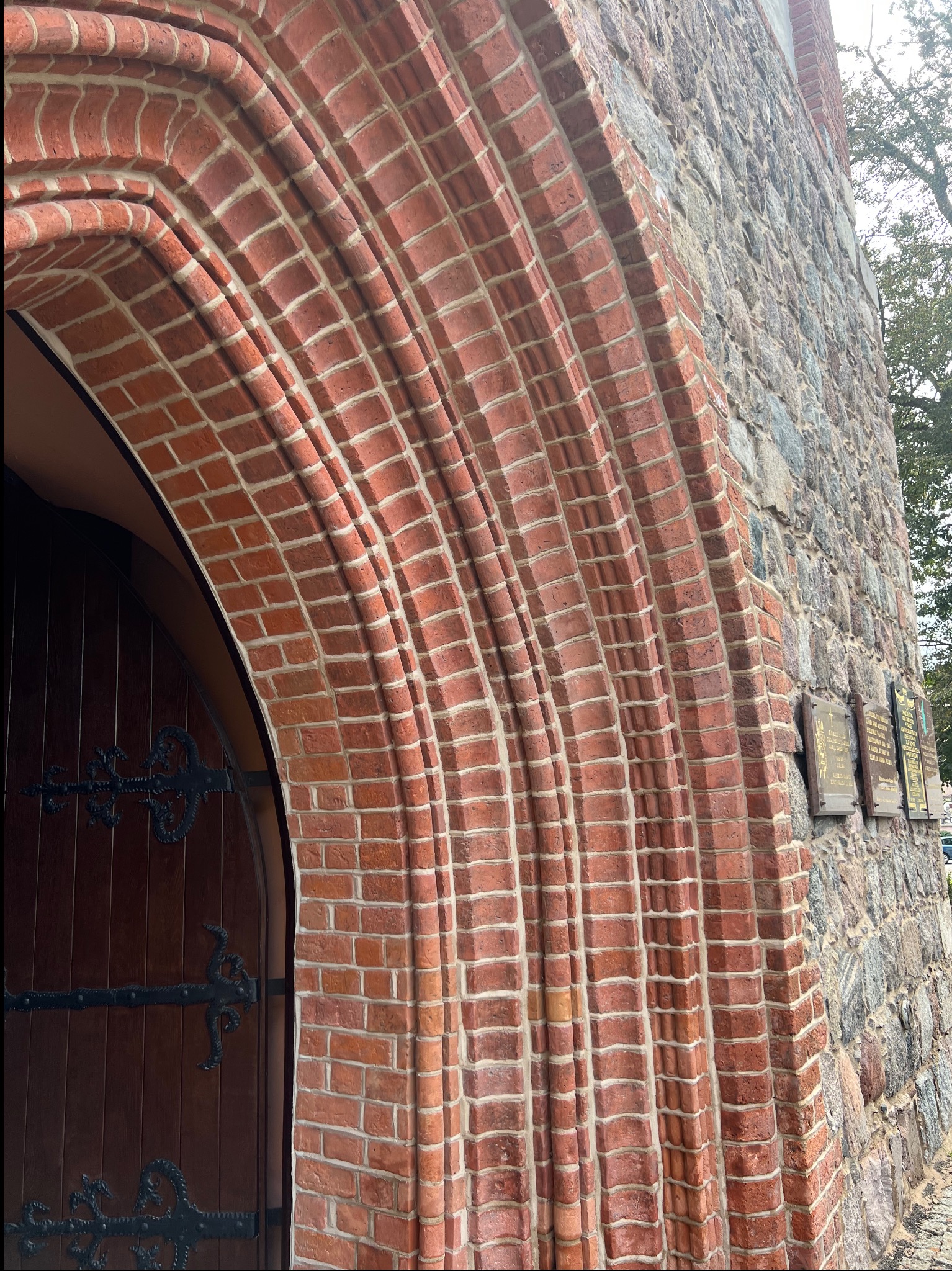 Ceglany łuk nad wejściem do zabytkowej kamienicy, z drewnianymi drzwiami i zdobieniami z kutego żelaza. Część kamiennej elewacji z tablicami pamiątkowymi.