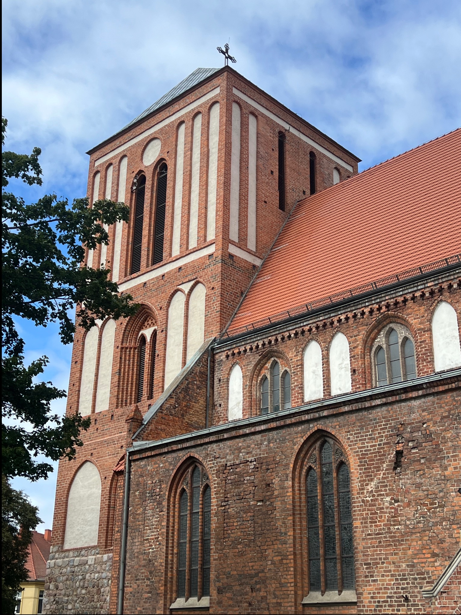 Gotycki kościół z cegły z wieżą i czerwonym dachem, widok z dołu, fragment drzewa po lewej. Charakterystyczne białe wstawki na wieży.